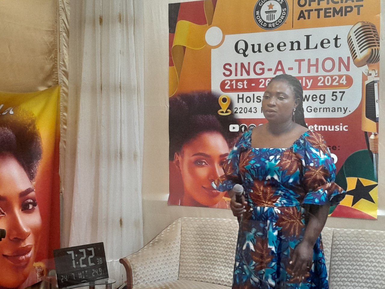 A woman stands singing into a microphone in a small room, beside a large printed poster announcing an official world record attempt with its dates