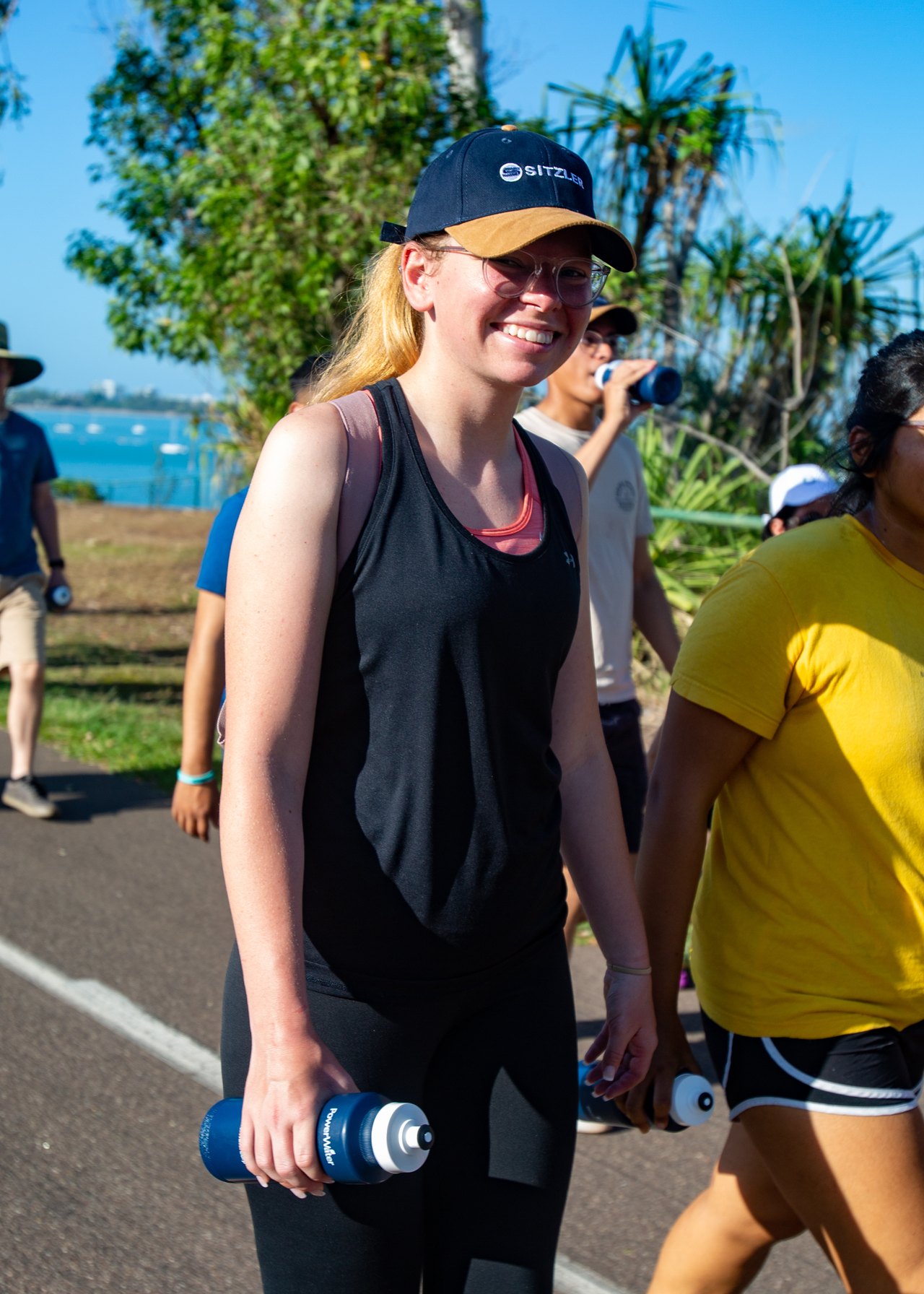 Volunteers in caps and casual clothes hand out bottles of water along a sunny roadside during a community event, smiling as people pass