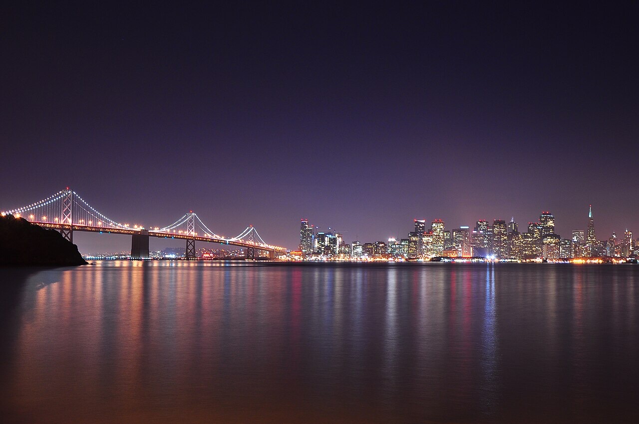 A city skyline glowing at night across dark water, a lit suspension bridge stretching in from the left