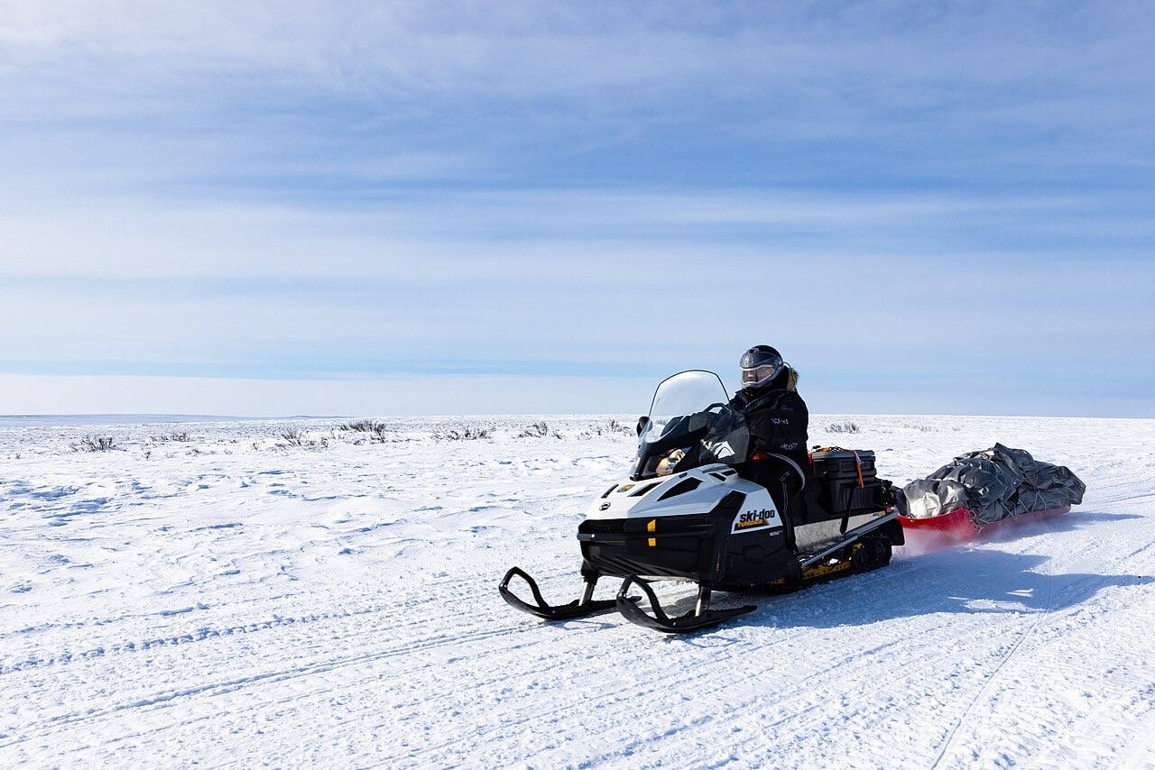 A rider in a helmet on a snowmachine pulling a loaded sled across flat white tundra under a blue sky
