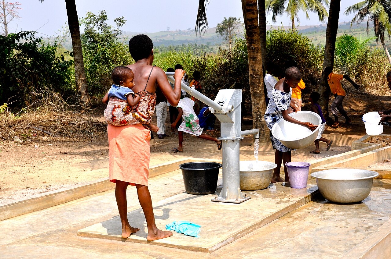 A woman with a baby on her back works the long handle of a hand pump on a concrete apron while water pours into a metal basin and children wait with buckets