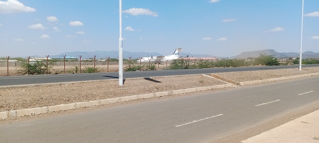 A white World Food Programme turboprop plane parked behind a wire fence at a dusty airstrip, with low buildings, thorn scrub and dry hills beyond