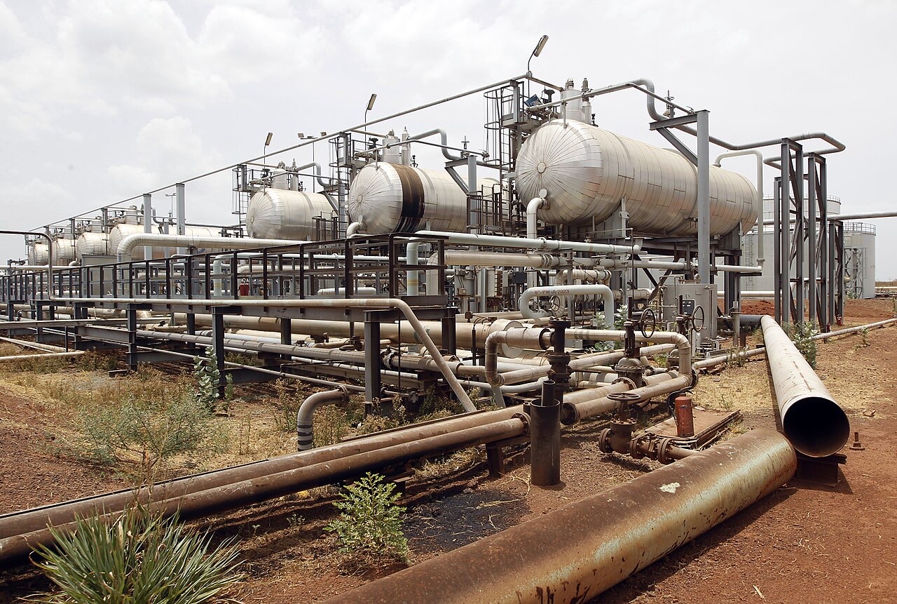 Rows of silver storage tanks and a web of gray and rust-brown pipes standing on red-brown earth under an overcast sky