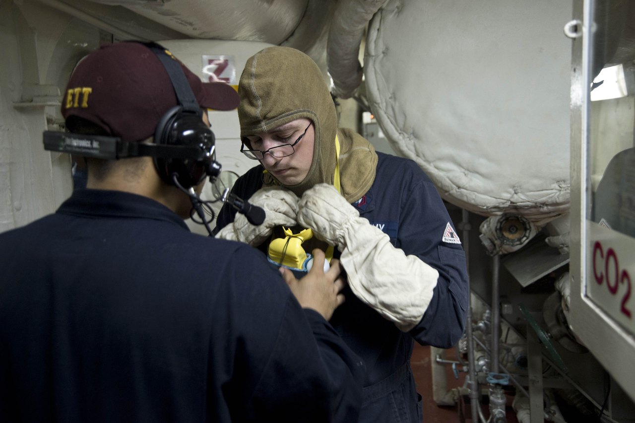 Two sailors in blue working uniforms practice a fire drill aboard a ship: one in a headset and cap helps the other pull on a protective hood, beside pipes and a red CO2 cabinet