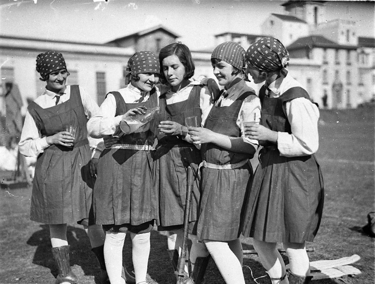 Black-and-white 1929 photograph of five schoolgirls in sports tunics and dark caps standing close together on a field, each holding a glass, smiling and drinking