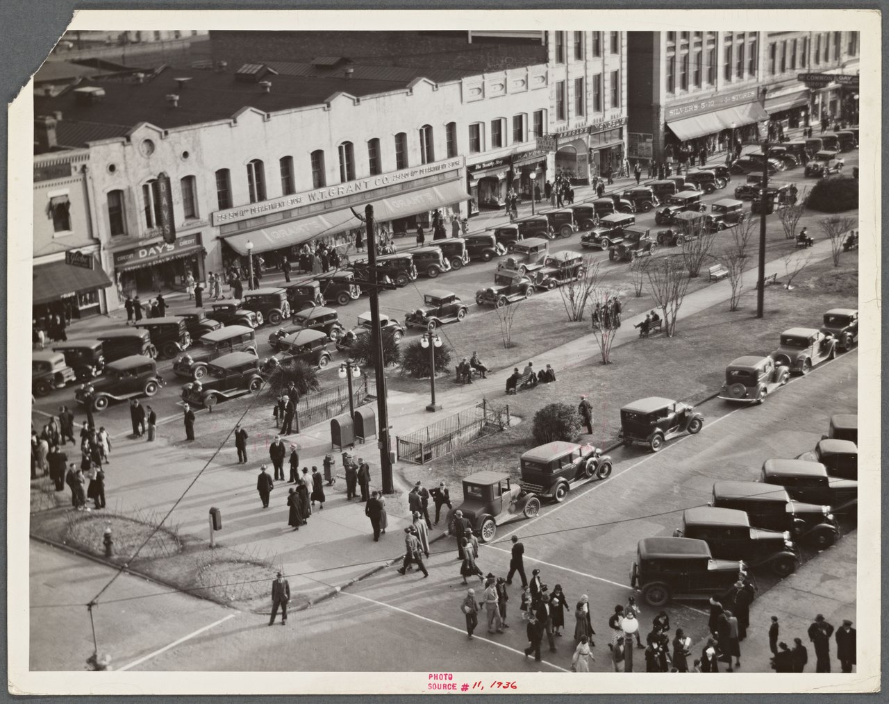 Black-and-white view from an upper window of a wide 1930s main street lined with shops and awnings, parked cars along both curbs, and dozens of pedestrians on the sidewalks and crossing