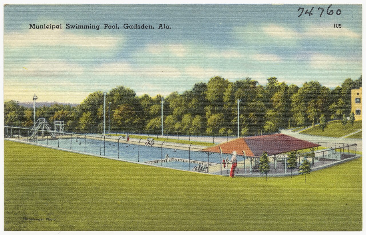 A colorful old postcard of a large outdoor municipal swimming pool with a red-roofed pavilion, lawns, trees, and a few swimmers, under a blue sky with white clouds