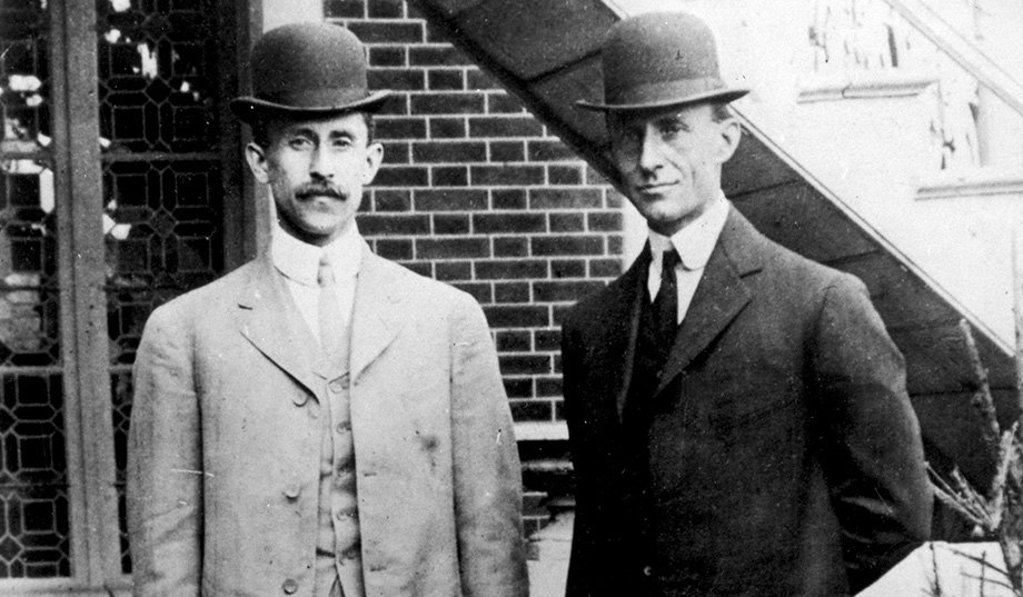 Black-and-white photograph of two men in dark suits and bowler hats standing side by side in front of a brick building, one with a mustache, both looking at the camera