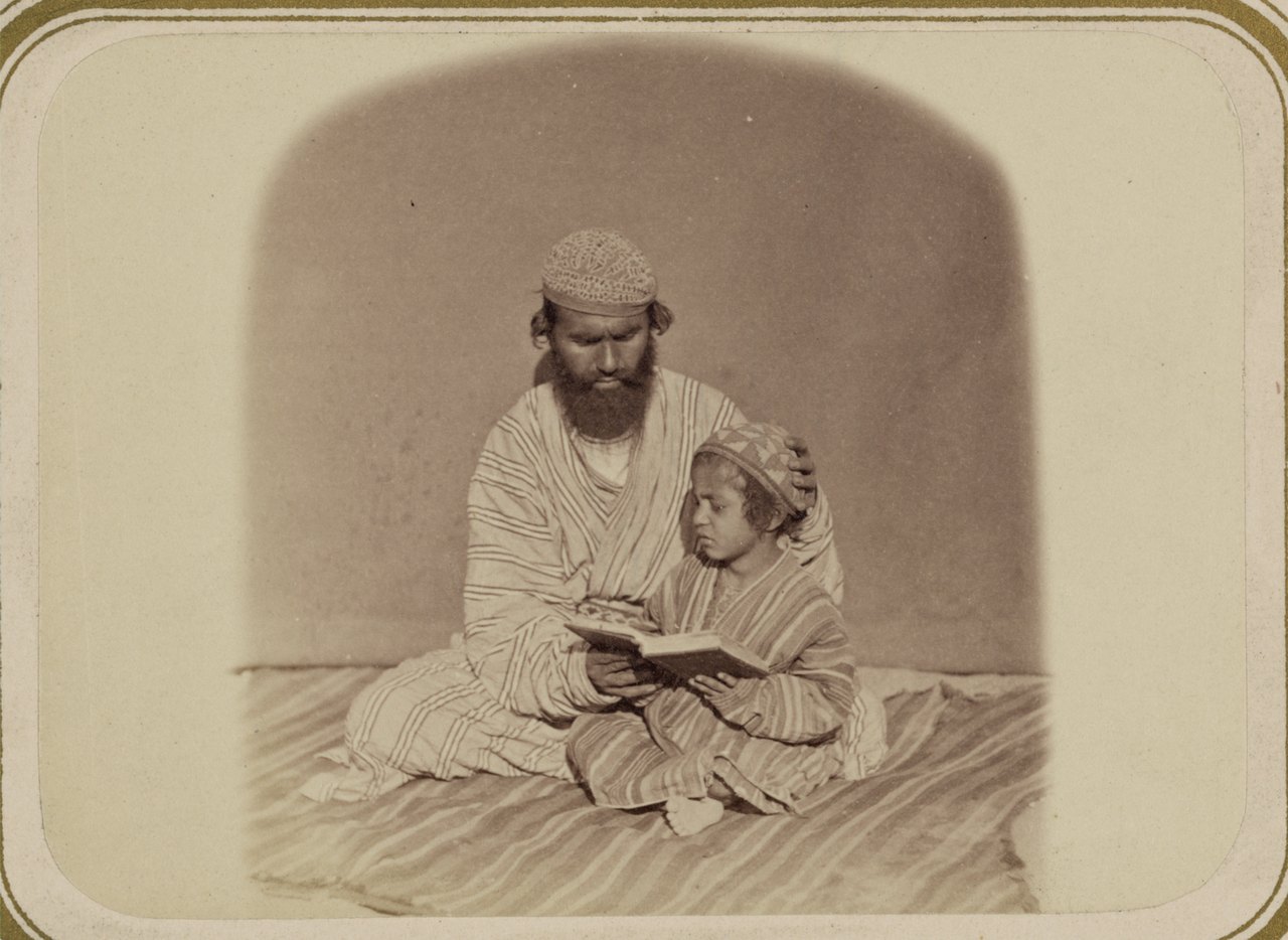 An old photograph of a seated man with his hand on the shoulder of a small child, who holds an open book and reads from it