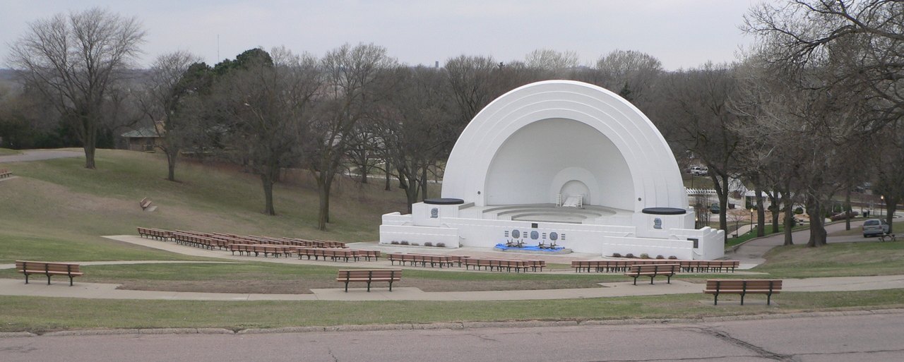 A white shell-shaped band stage in a public park, with rows of empty benches on the grass in front of it and bare trees behind