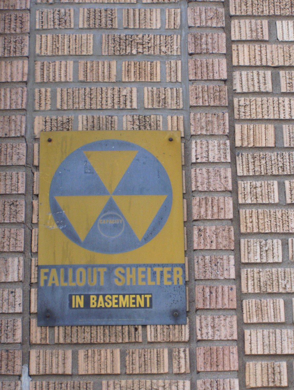 A yellow and black fallout shelter sign bolted to a brick wall, reading FALLOUT SHELTER IN BASEMENT