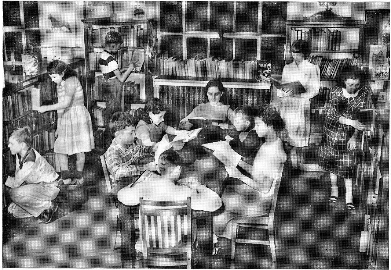 A black-and-white photograph of a busy indoor library room: children pulling books from low shelves that run around the walls, and others sitting at a round table absorbed in reading