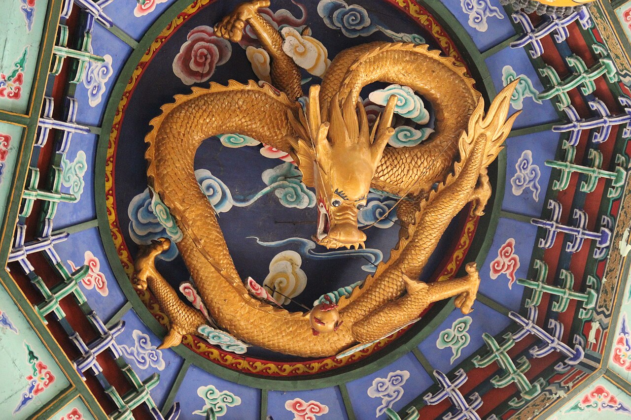 A gilded carved dragon coiled inside a round painted ceiling panel, its long snake-like body ringed by blue and green clouds, with whiskers and four clawed feet