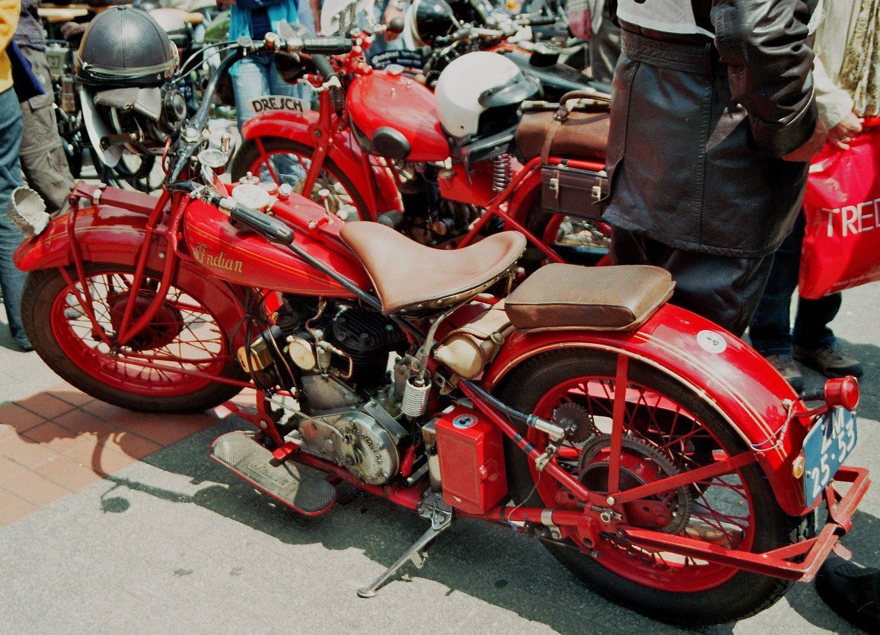 A bright red vintage motorcycle with a brown leather saddle, exposed engine, and spoked wheels stands on a street at a rally, with people and other motorcycles behind it