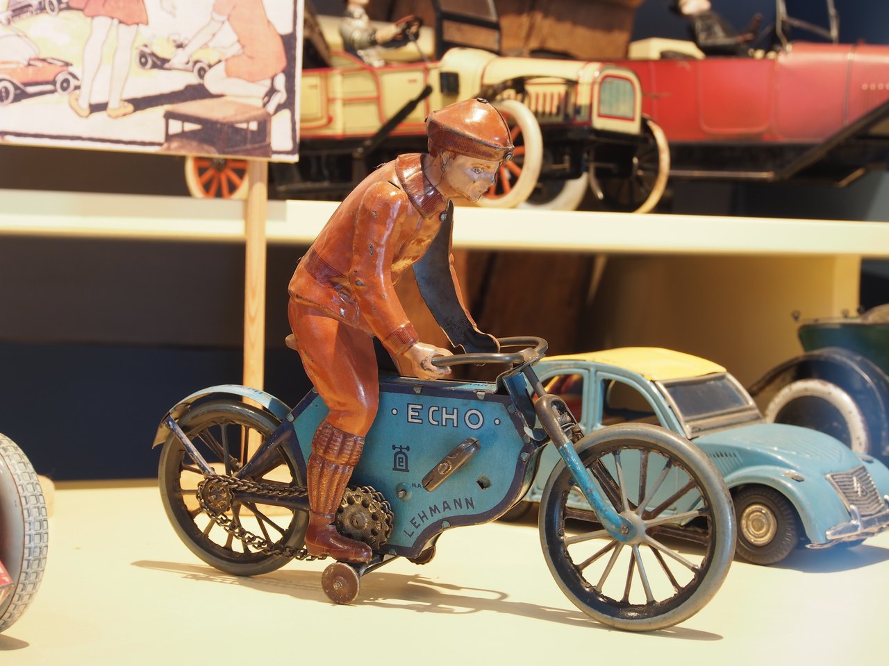 A painted tin toy of a rider in an orange suit and cap on a small blue motorcycle with spoked wheels, displayed on a museum shelf with other old tin vehicles behind it