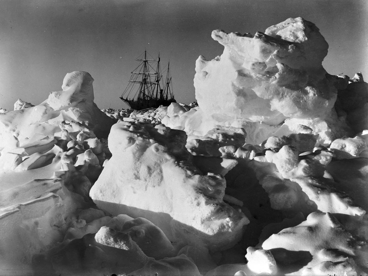 Black-and-white photograph of a three-masted wooden ship caught in a field of broken, heaped-up sea ice, with huge tilted blocks of ice in the foreground