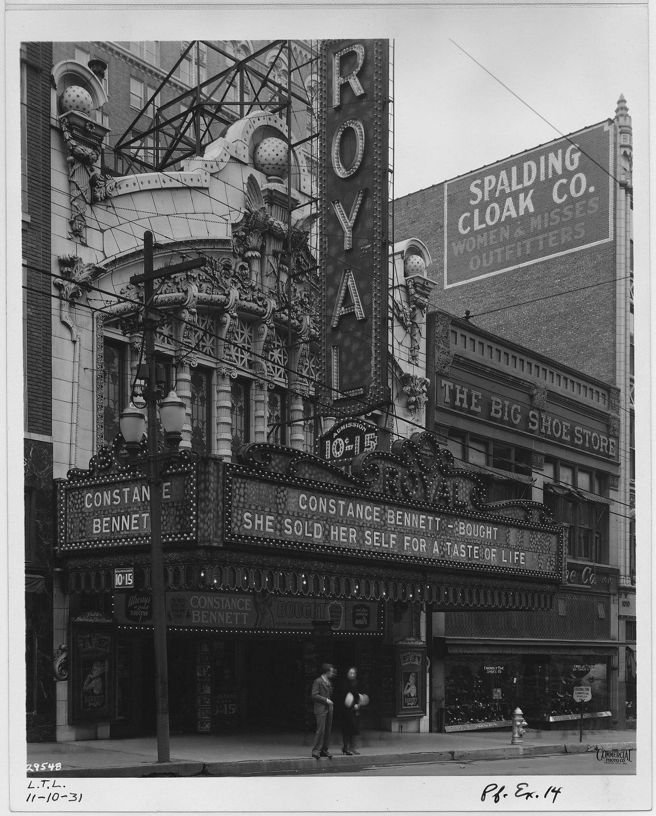 Black-and-white 1931 street photograph of an ornate theater front with a tall vertical sign reading ROYAL, a marquee of light bulbs advertising a Constance Bennett film, and two people walking past