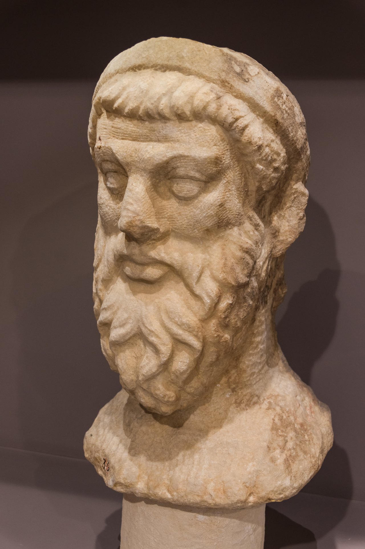 A bearded head carved from pale marble, the hair and beard cut into deep curling grooves while parts of the surface are still rough