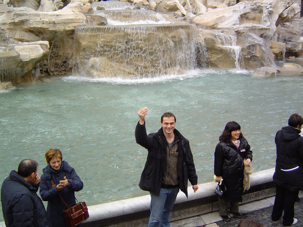 Visitors standing at the low wall of a large fountain, one with an arm raised mid-throw, the pale green water and its stone basin filling the frame