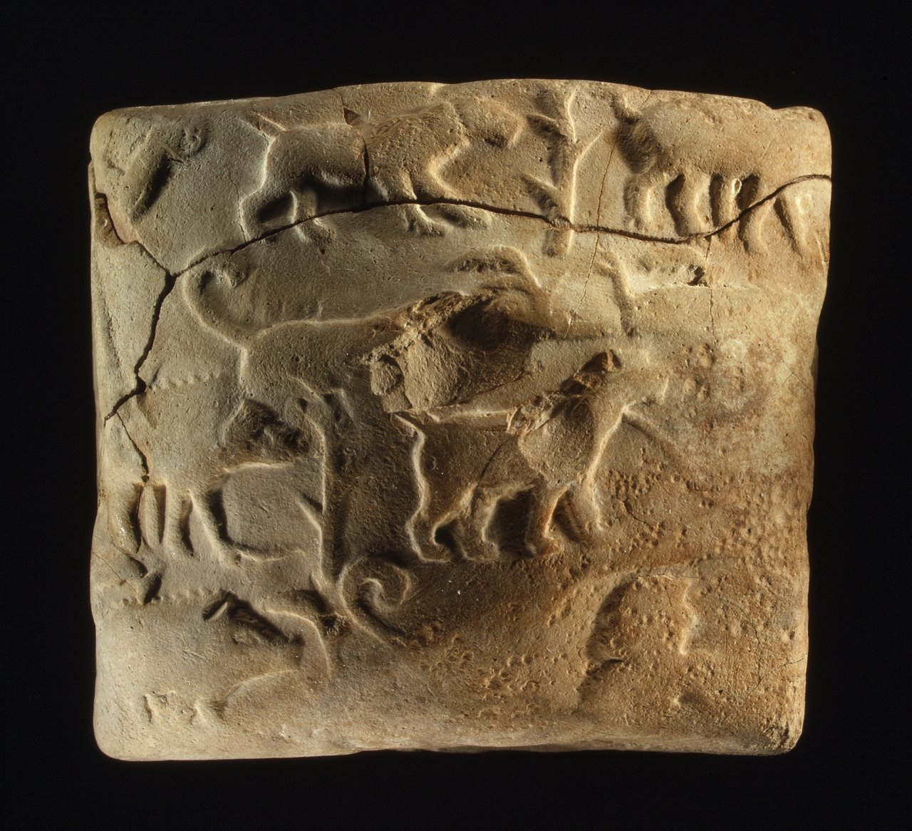 A small cracked clay tablet whose surface carries rolled-in figures of dogs and boars, raised and recessed, alongside wedge-shaped writing