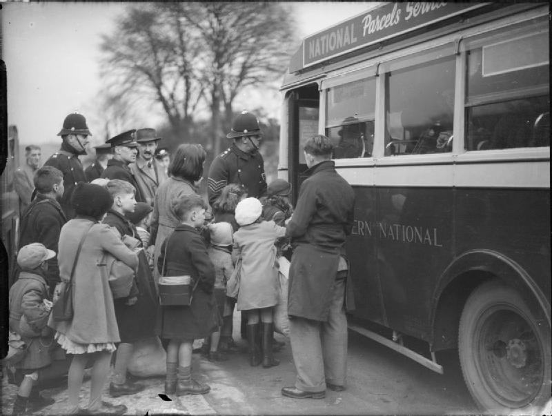 A black-and-white photograph of a crowd of small children in coats, carrying bags and boxes, being helped by policemen onto a single-deck bus with National painted on its side