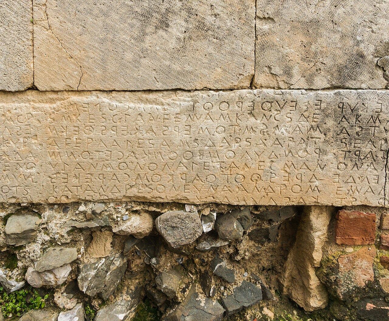 Rows of ancient Greek letters cut into large pale stone blocks in a wall, with rough rubble stonework beneath them