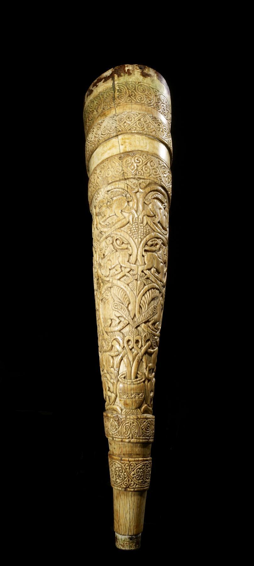 A tall, tapering horn of pale carved ivory photographed against black, its surface covered in animals climbing through scrolling vines between bands of pattern
