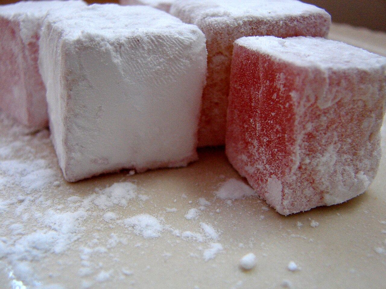 A close-up of soft pink cubes of Turkish delight thickly dusted with white powdered sugar, with loose sugar scattered on the plate around them