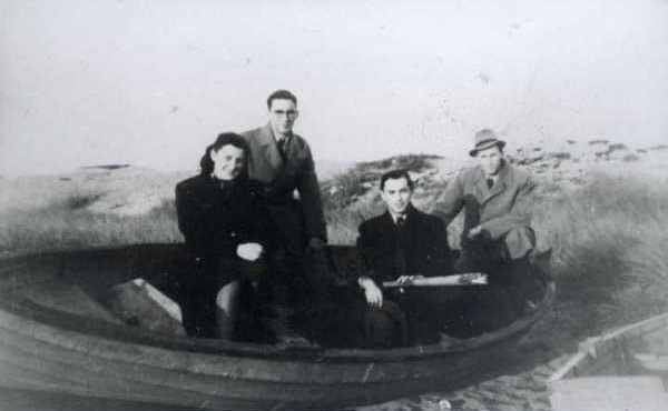 A grainy black-and-white photograph of four people, a woman and three men in coats, sitting and standing in a wooden rowboat pulled up on a grassy shore, one of them holding an oar