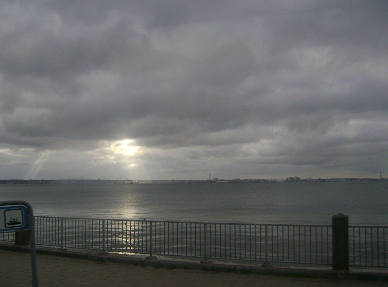 A gray, overcast view from a railed waterfront promenade across a stretch of sea to a low far shore with buildings and chimneys, sunlight breaking through the clouds onto the water
