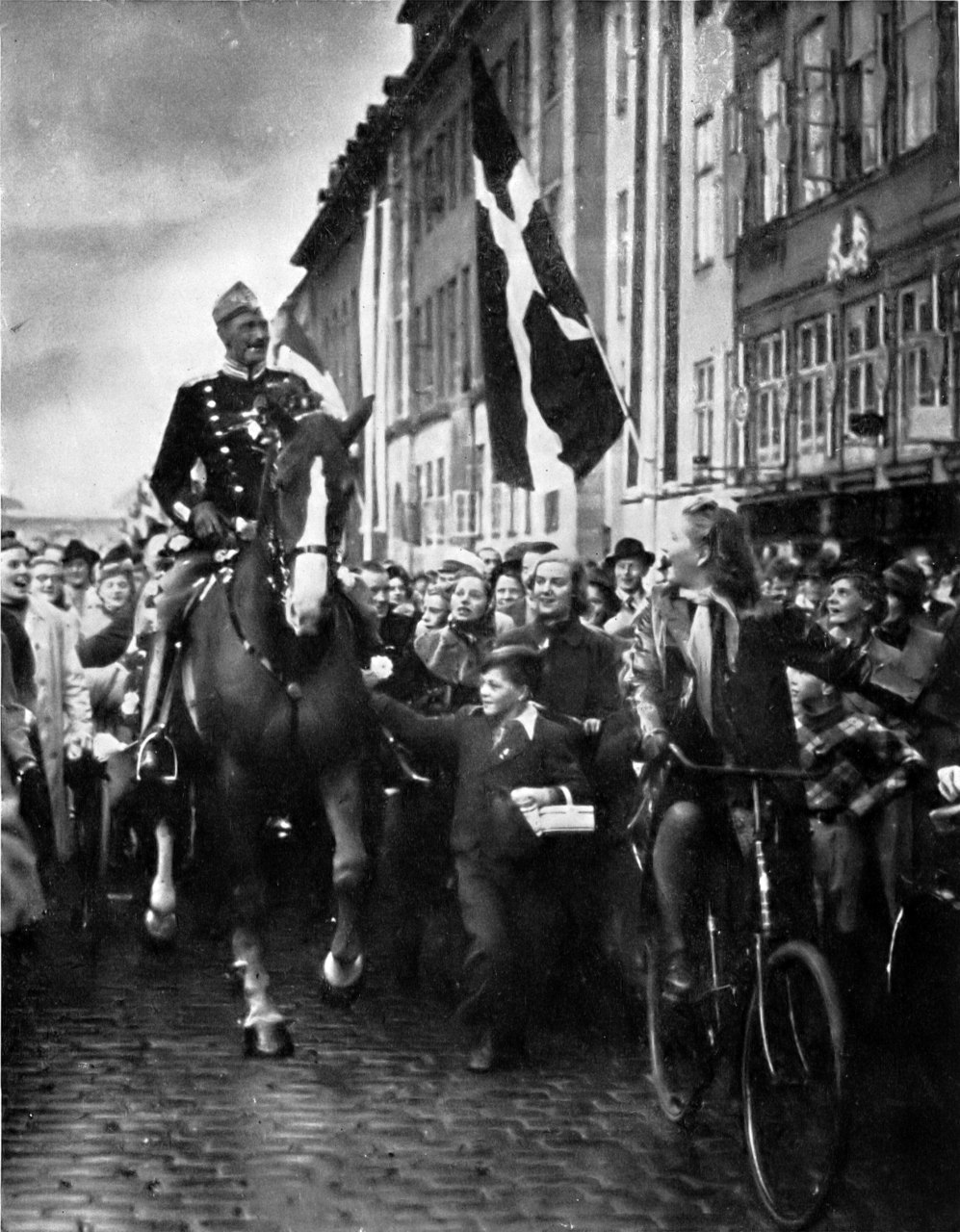 A black-and-white photograph of a mustached king in a dark uniform riding a horse through a cobbled Copenhagen street, surrounded by a dense crowd, with Danish flags hanging from the buildings