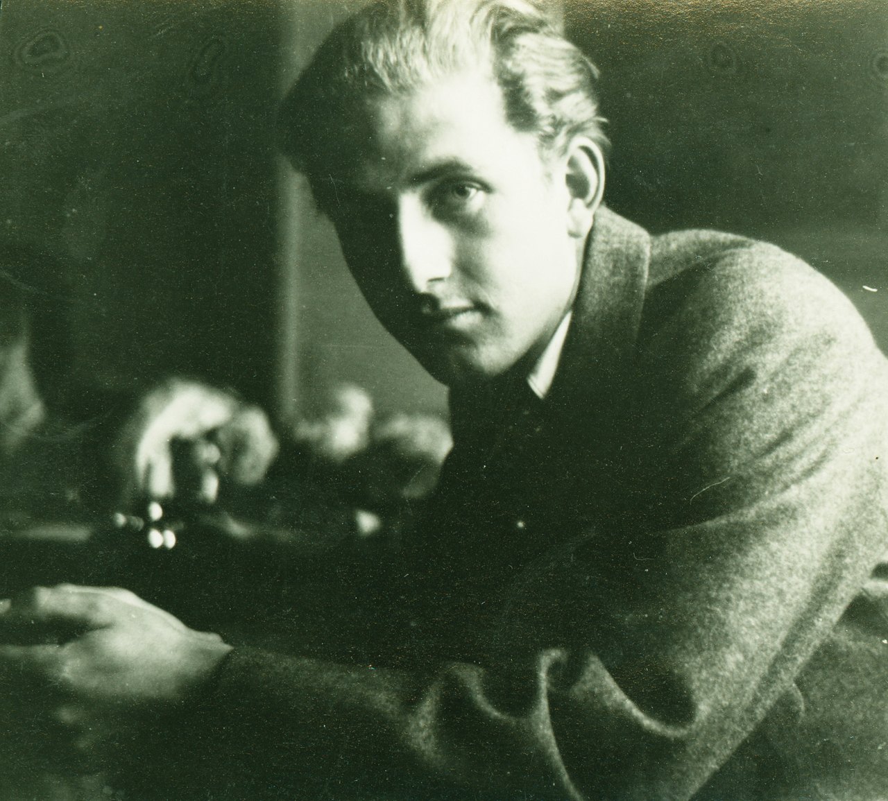 A sepia-toned portrait photograph of a young man with swept-back fair hair, leaning forward on his arms in a tweed jacket and looking directly at the camera