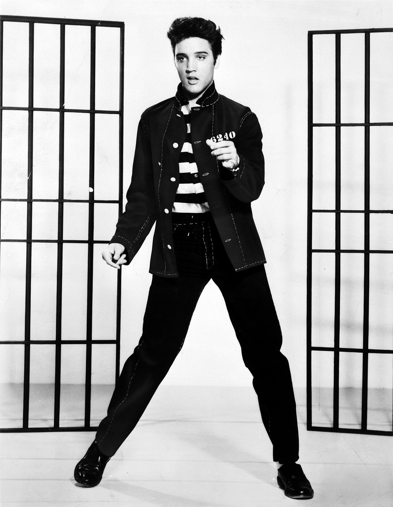 A black-and-white full-length publicity photograph of Elvis Presley in a dark denim jacket, caught mid-dance-step in front of stylized jail bars, his dark hair swept up and back in a high greased pompadour