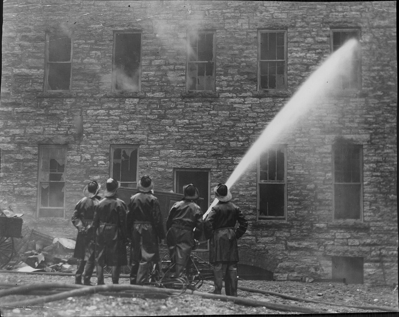 A black-and-white photograph of five helmeted firefighters in long coats aiming one hose stream through an upper window of a stone building, with smoke drifting from several broken upper windows