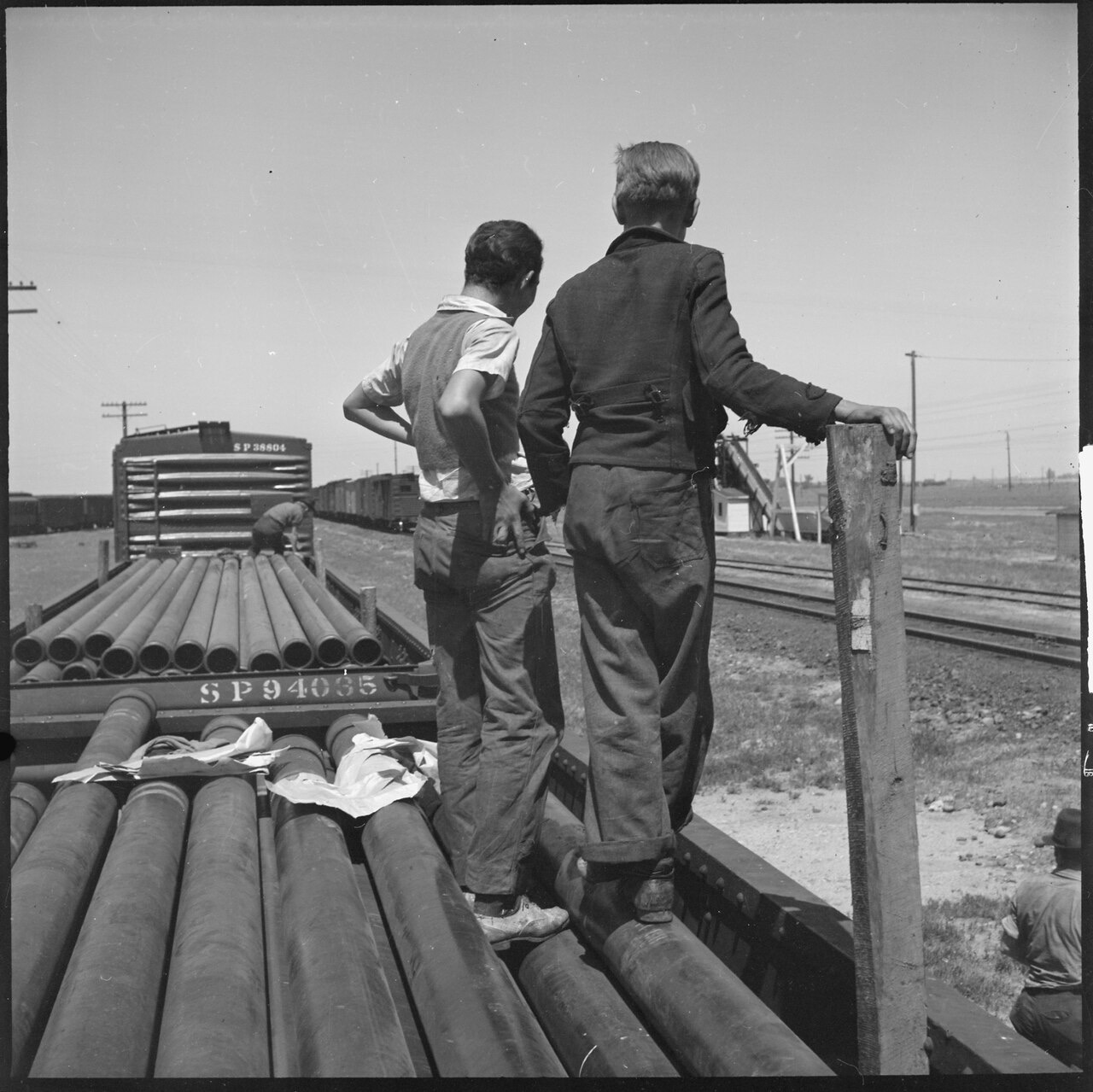 A black-and-white photograph of two boys in work clothes standing on a flatcar loaded with steel pipe, looking down a sunlit rail yard, one steadying himself against a wooden post