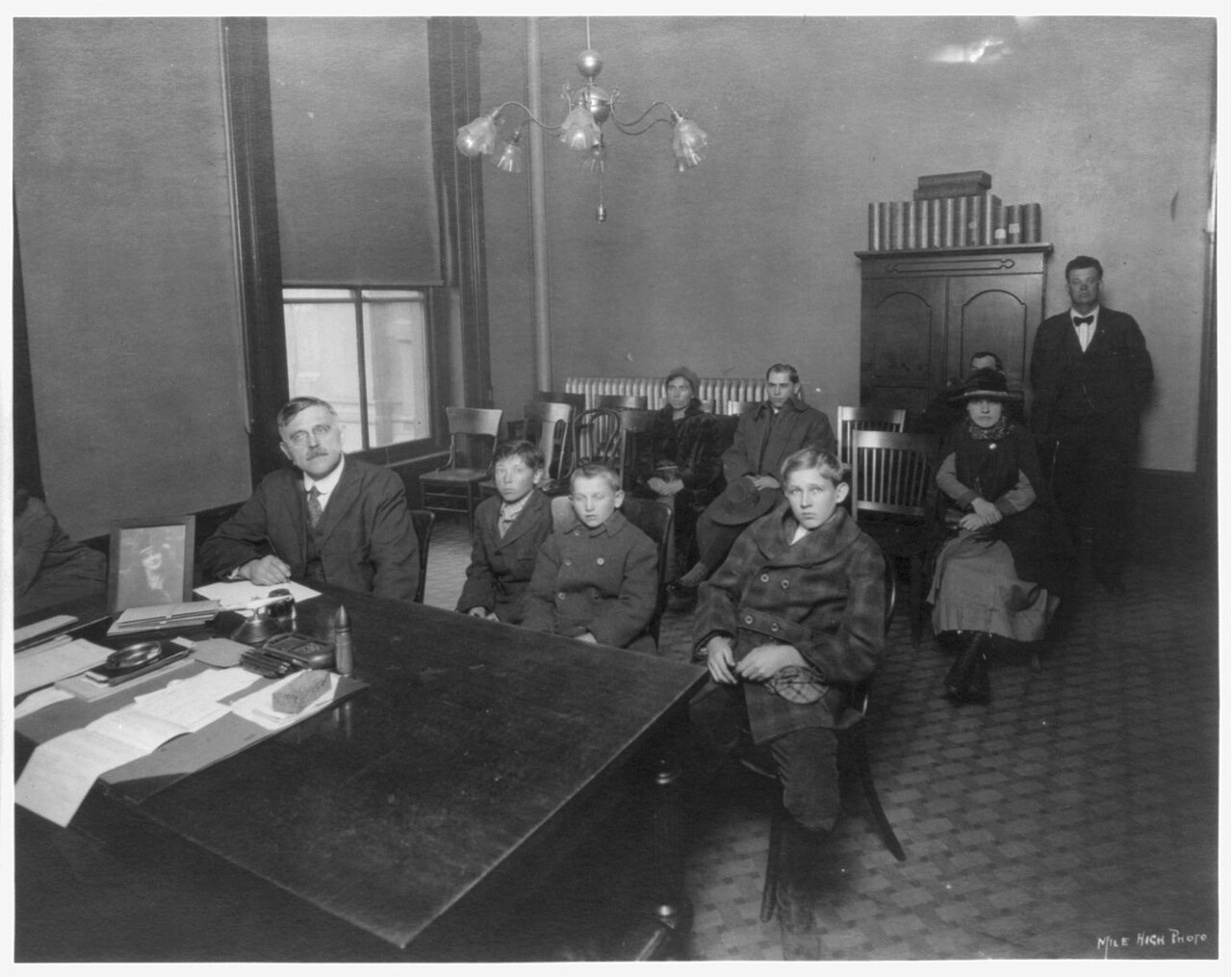 A black-and-white photograph of a paneled office where a mustached judge sits at a large document-covered desk; three boys sit in a row of chairs beside the desk, with several adults seated and standing behind them