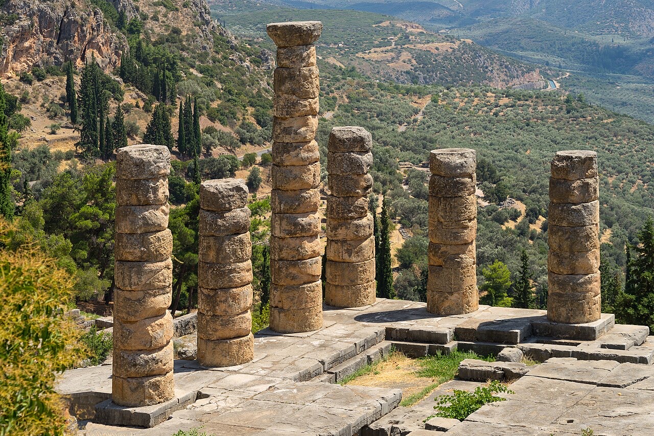 Six weathered stone columns of a ruined Greek temple standing on a stone platform, high above a valley of olive trees and wooded hills