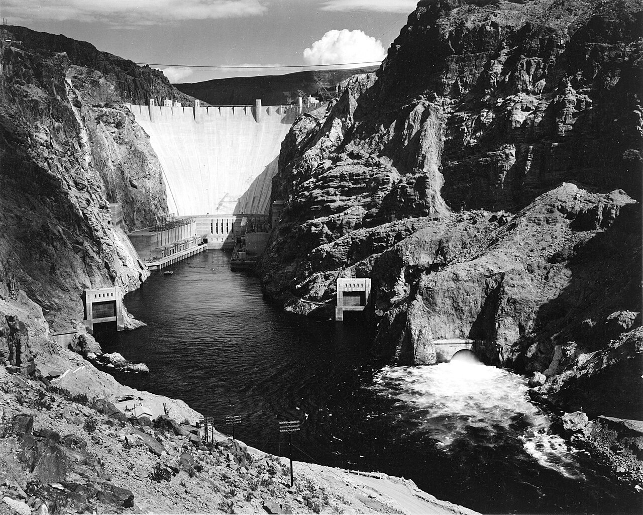 A black-and-white view down a rocky canyon: a massive curved concrete dam spans the gorge, with a white plume of water jetting into the dark river below