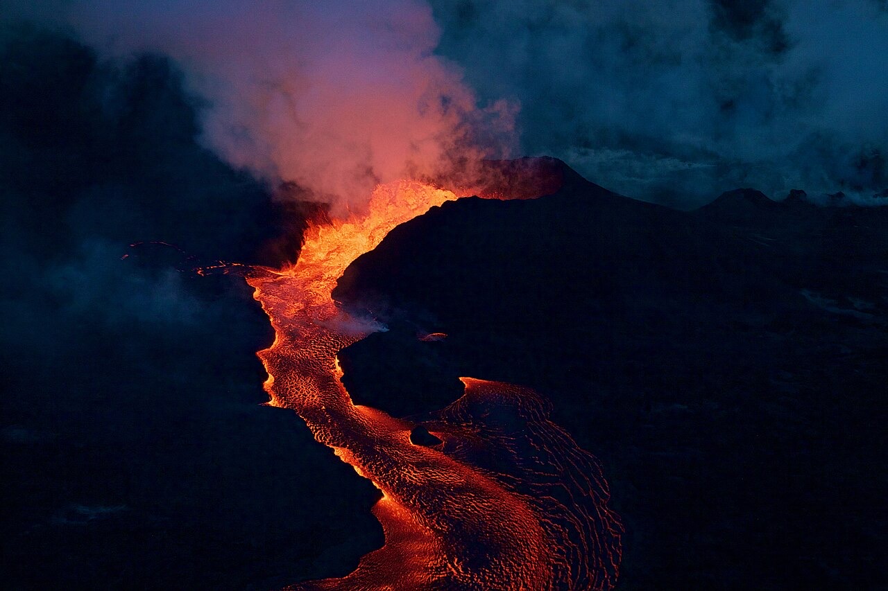 A glowing orange river of lava pours from a dark erupting cone at dusk, its channel snaking toward the foreground under clouds of steam
