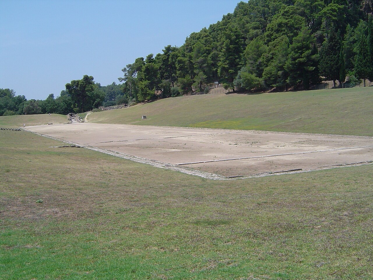 A long rectangular strip of bare pale clay stretches across a grassy hollow ringed by low green banks, with pine-covered hills behind