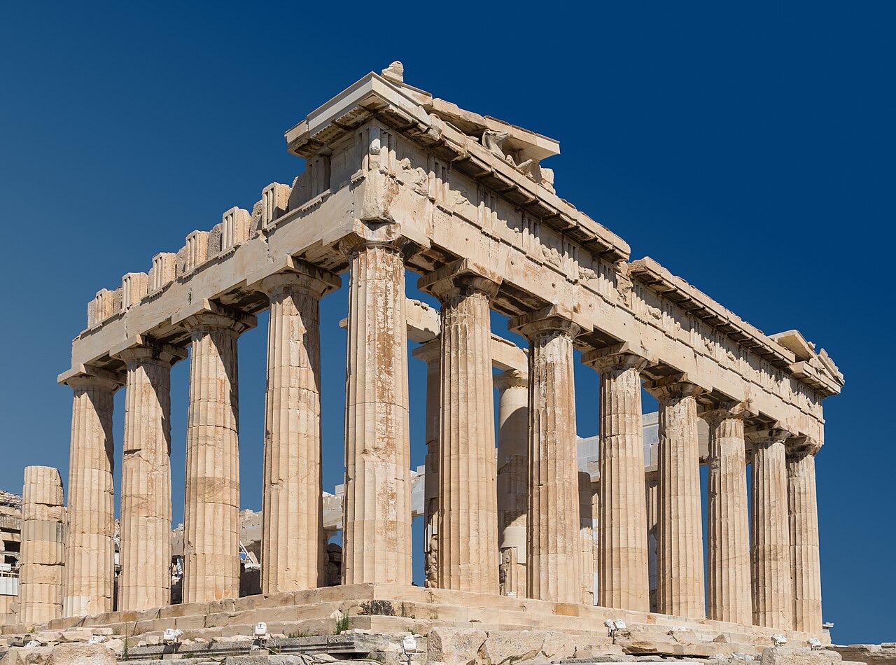 The Parthenon seen from a corner under a deep blue sky: weathered marble Doric columns supporting a broken entablature and pediment