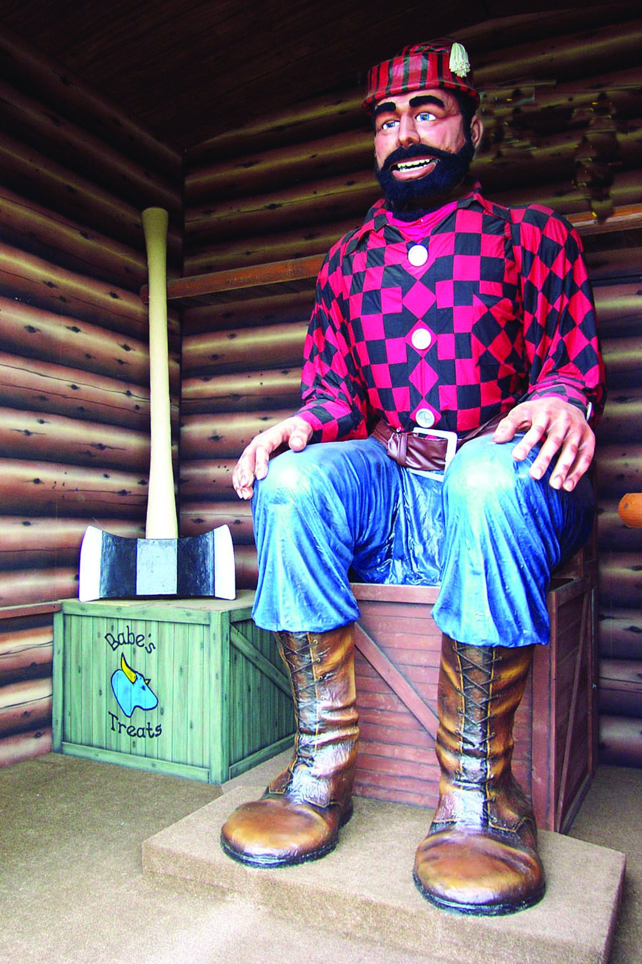 A giant painted statue of a bearded lumberjack in a red-and-black checked shirt, blue trousers and boots sits on a log bench inside a wooden building, dwarfing the doorway beside it