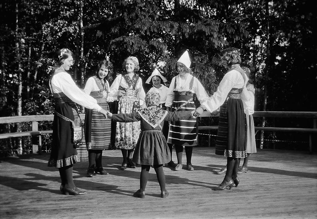 Black-and-white photograph of young women and girls in Swedish folk costumes, white blouses, dark skirts and pointed caps, dancing in a ring on an outdoor wooden floor among trees