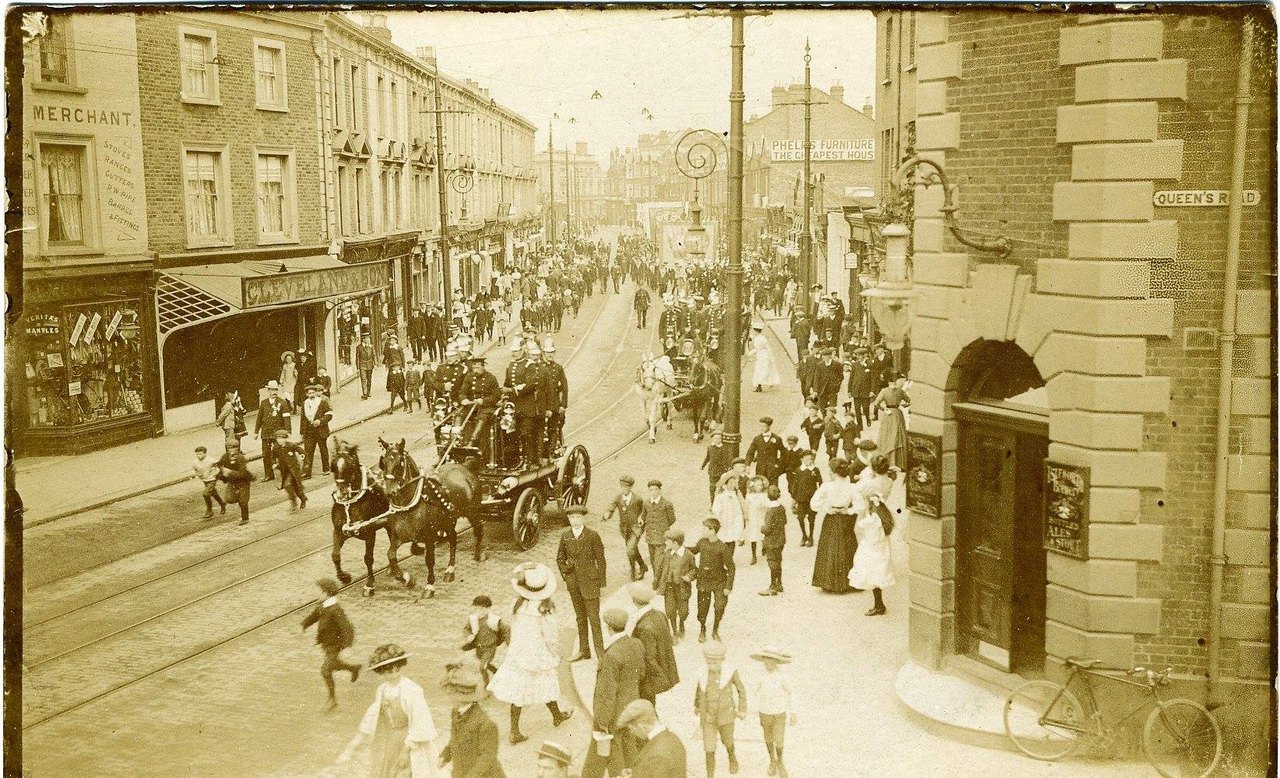 Sepia postcard photograph of a town street in the early 1900s: firemen in shining helmets ride a horse-drawn fire engine down the middle of the road while crowds of people in hats and long dresses line both sides