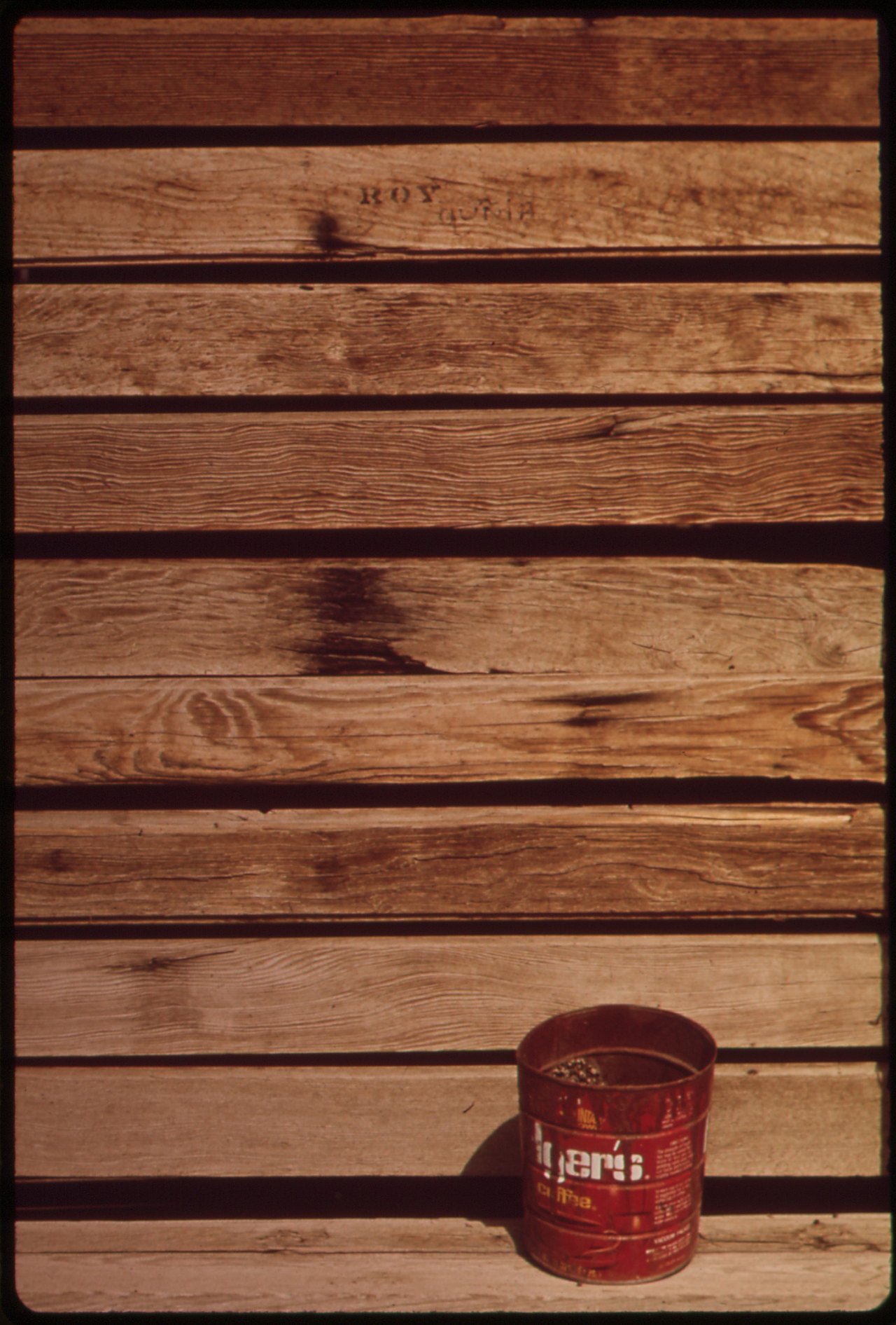 A single rusted red coffee can stands on the ground against a wall of weathered horizontal wooden planks