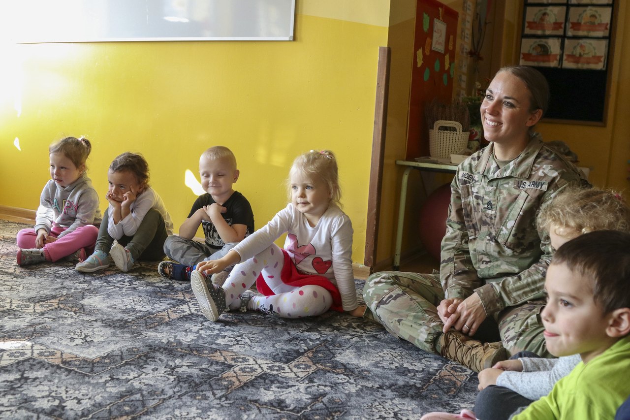 Young children sitting cross-legged on the floor in a bright classroom, listening to an adult seated with them