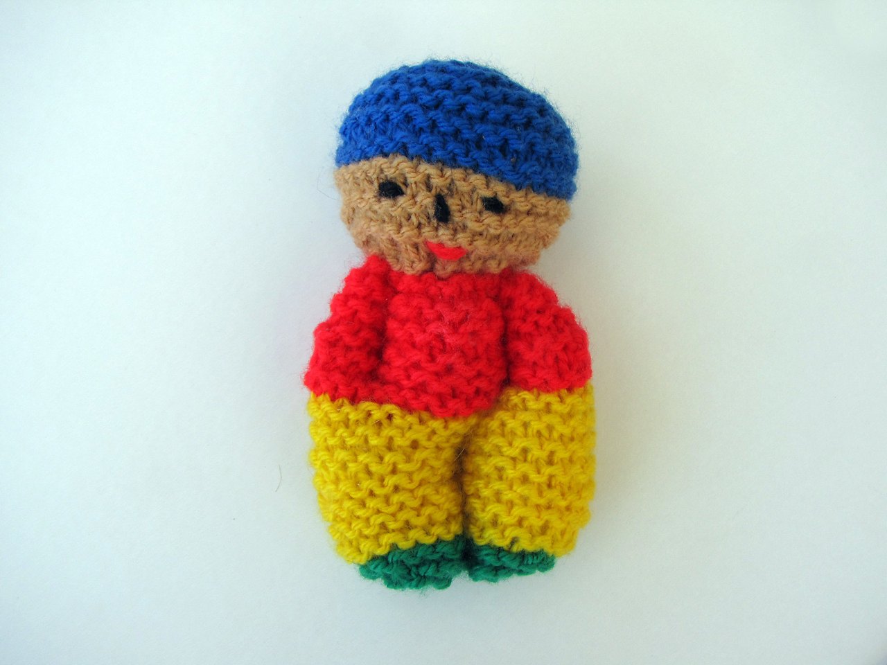 A small knitted rag doll in red, yellow, green and blue wool, with a plain stitched face and a round blue cap, lying on a pale surface