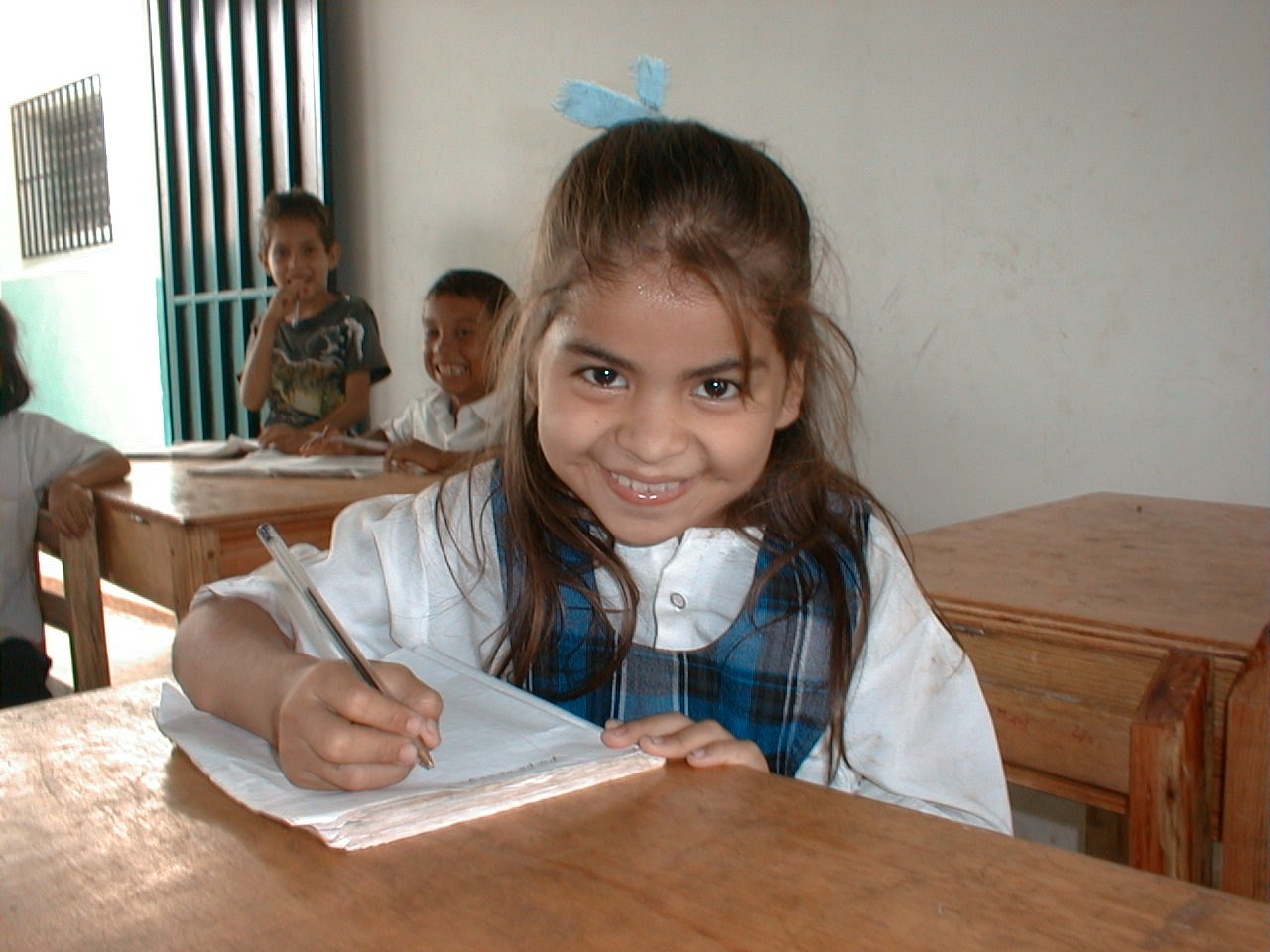 A young girl at a wooden school desk smiles at the camera while writing in an open exercise book, holding her pencil carefully, with classmates behind her