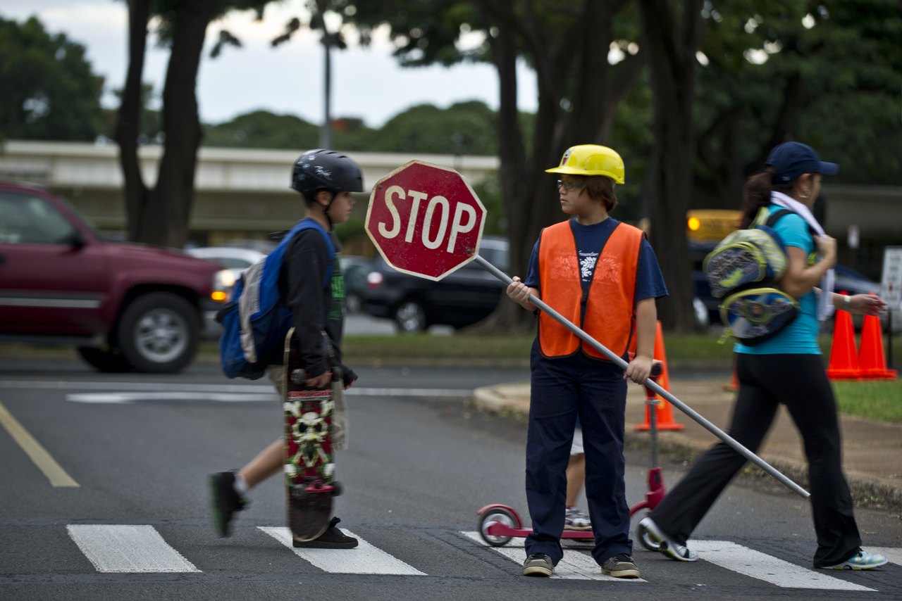 A child in an orange safety vest holds up a red stop sign at a marked crosswalk while other children cross the road with backpacks and scooters