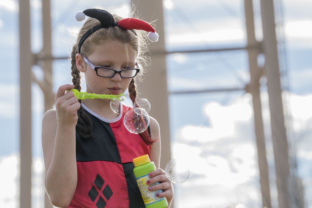 A young girl in a black-and-red Halloween costume with a horned headband and glasses stands indoors holding a small toy, looking sideways at the camera